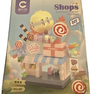 May include: A building block set box featuring a colorful candy shop model with a blue and white striped awning, a large yellow sphere, and lollipop decorations. The packaging displays the text Shops, Sweet Shop, Candy Shop, and PCS.289.