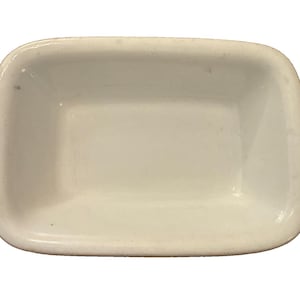 May include: A small, rectangular white ceramic dish with rounded corners and a smooth, glossy finish. The dish features a deep, open basin with slightly sloped interior walls and a flat base, presenting a clean and simple design for holding small items.