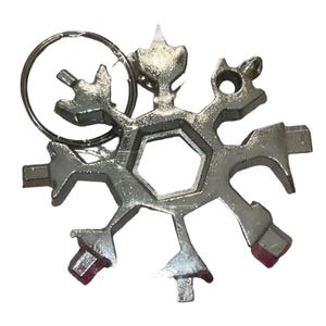 Könnte beinhalten: Ein silbernes Multi-Tool in Form eines Schneeflocks, mit verschiedenen integrierten Werkzeugen. Es enthält einen Schlüsselring und eine zentrale sechseckige Öffnung. Das Werkzeug hat eine glänzende, metallische Oberfläche und ist für mehrere Zwecke konzipiert.