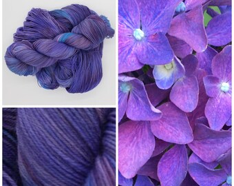 May include: A skein of purple and blue yarn with a variegated colour pattern. The yarn is arranged in a loose pile. The image also shows a close-up of the yarn and a cluster of purple hydrangea flowers.