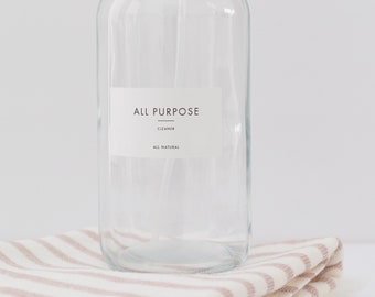 Pode incluir: Um frasco de spray de vidro transparente com um bico de pulverização branco e um rótulo branco que diz "ALL PURPOSE CLEANER ALL NATURAL". O frasco está sobre uma toalha de listras rosa e brancas dobrada.