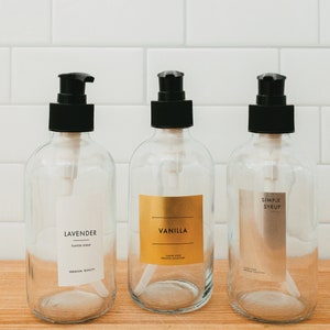 Puede incluir: Tres botellas de vidrio transparente con tapas de bomba negras y etiquetas. Las etiquetas dicen "Jarabe de sabor a lavanda", "Jarabe de sabor a vainilla" y "Jarabe simple".