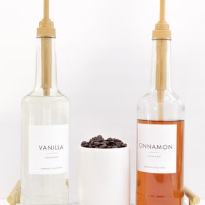 May include: Two glass bottles with pumps filled with clear and brown liquids. The bottles have labels that say "Vanilla Flavor Syrup Premium Collection" and "Cinnamon Flavor Syrup Premium Collection". A white ceramic cup filled with coffee beans sits between the bottles. The bottles and cup are on a white marble tray with gold metal handles.