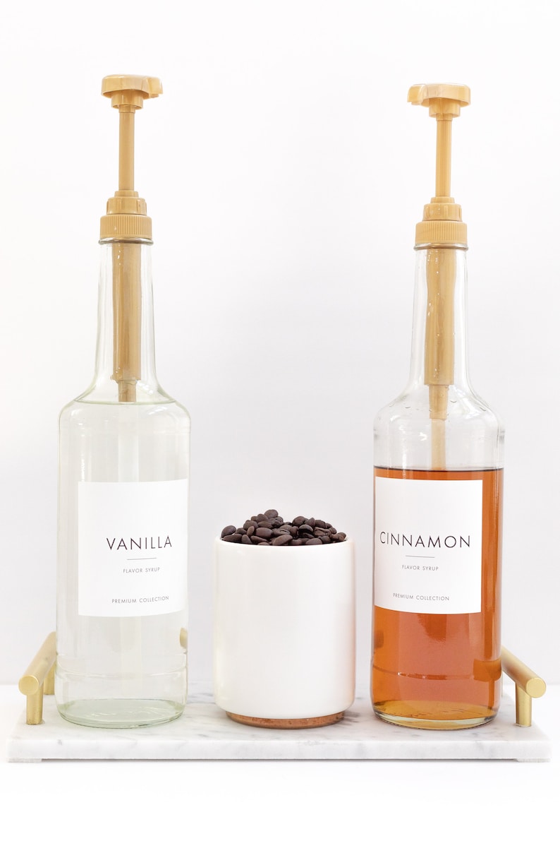 May include: Two glass bottles with pumps filled with clear and brown liquids. The bottles have labels that say "Vanilla Flavor Syrup Premium Collection" and "Cinnamon Flavor Syrup Premium Collection". A white ceramic cup filled with coffee beans sits between the bottles. The bottles and cup are on a white marble tray with gold metal handles.