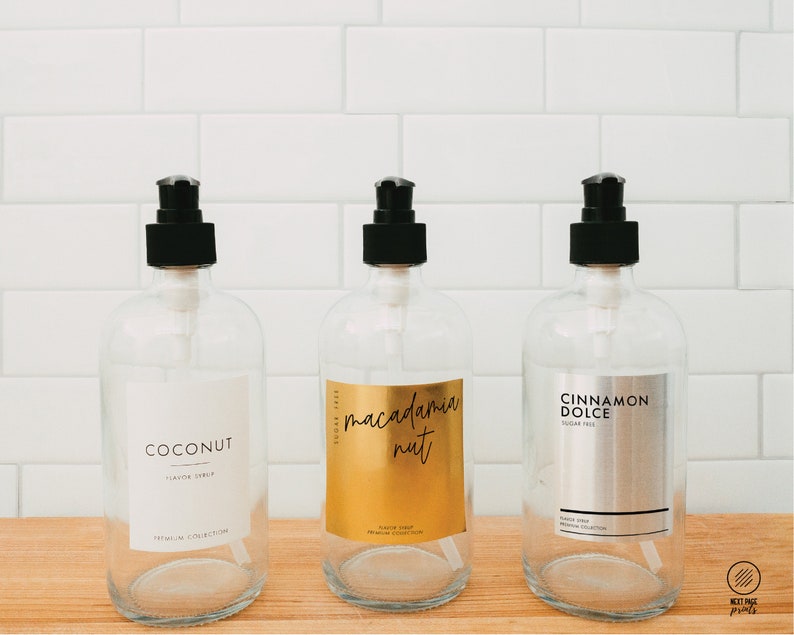 Puede incluir: Tres botellas de vidrio transparente con tapas de bomba negras y etiquetas. Las etiquetas son doradas, plateadas y blancas con el texto "Coconut", "Macadamia Nut" y "Cinnamon Dolce".