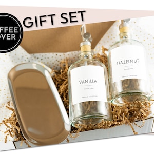 May include: Gift set for coffee lovers with two glass bottles of flavored syrup, one labeled "Vanilla" and the other "Hazelnut", and a silver tray. The bottles have a pump dispenser and are sitting on brown shredded paper in a white box.