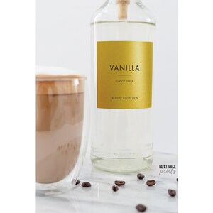 May include: A clear glass bottle with a gold label that says "Vanilla Flavored Syrup Premium Collection". The bottle is sitting on a white surface with coffee beans scattered around it. There is a glass of coffee with a layer of foam on top in the background.