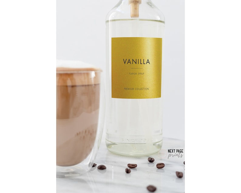 May include: A clear glass bottle with a gold label that says "Vanilla Flavored Syrup Premium Collection". The bottle is sitting on a white surface with coffee beans scattered around it. There is a glass of coffee with a layer of foam on top in the background.