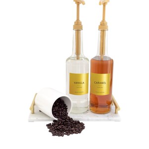 May include: Two glass bottles with pumps, one filled with clear liquid and labeled "Vanilla", the other filled with brown liquid and labeled "Caramel". A white ceramic cup with coffee beans spills out onto a white marble surface.