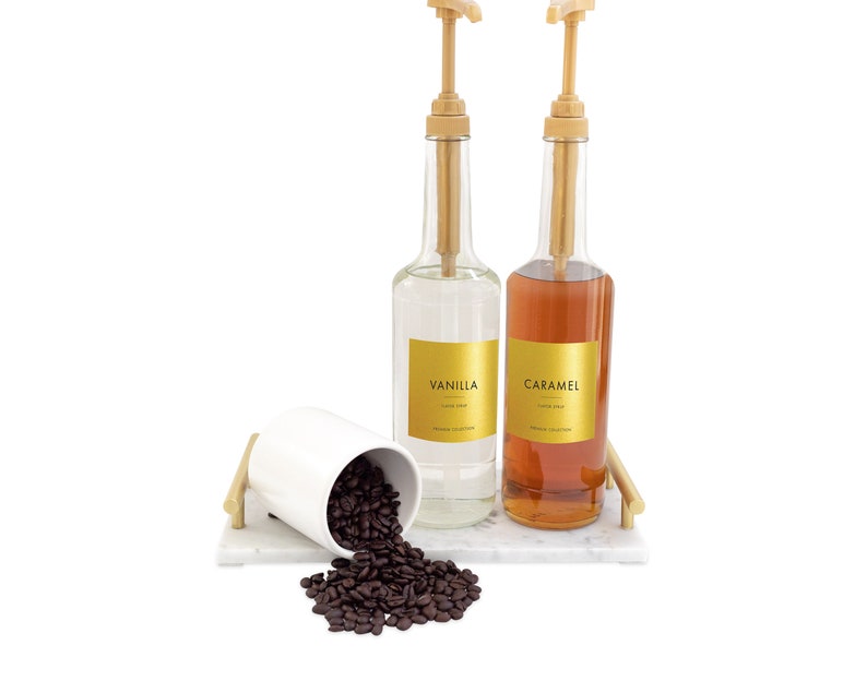 May include: Two glass bottles with pumps, one filled with clear liquid and labeled "Vanilla", the other filled with brown liquid and labeled "Caramel". A white ceramic cup with coffee beans spills out onto a white marble surface.