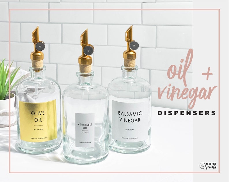 May include: Three glass bottles with gold tops and labels. The labels read "Olive Oil", "Vegetable Oil", and "Balsamic Vinegar". The bottles are on a white countertop with a white tiled backsplash. The text "oil + vinegar dispensers" is in the upper right corner of the image.