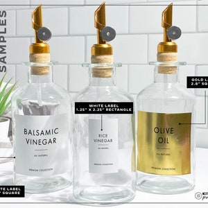 Puede incluir: Tres botellas de vidrio transparente con boquillas doradas y tapones de corcho. Cada botella tiene una etiqueta: Vinagre Bals&aacute;mico, Vinagre de Arroz y Aceite de Oliva. Las etiquetas son blancas o doradas con texto negro.