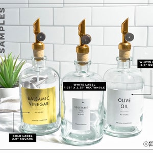 May include: Three glass bottles with gold, white, and gold labels. The bottles are filled with liquids and have gold and black pump tops. The labels have text that reads "Balsamic Vinegar", "Vegetable Oil", and "Olive Oil". The labels also include the size and shape of the label.