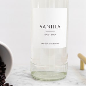 May include: A clear glass bottle with a white label that reads "Vanilla Flavor Syrup Premium Collection". The bottle is sitting on a white marble surface.