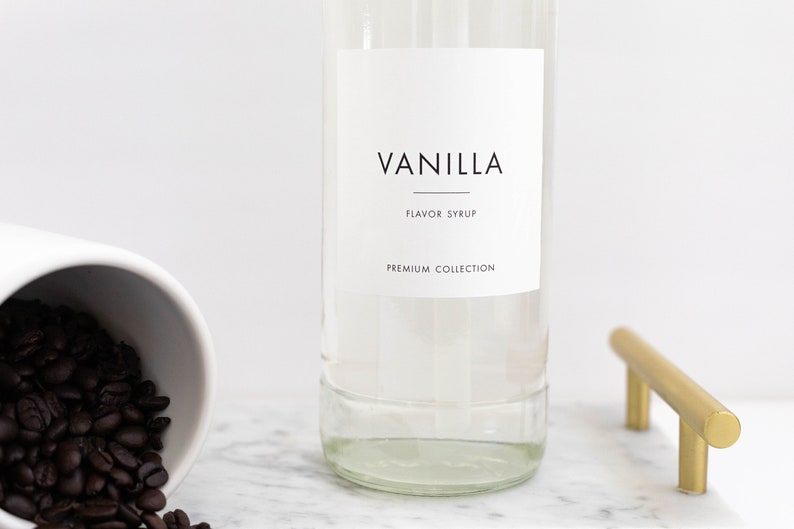 May include: A clear glass bottle with a white label that reads "Vanilla Flavor Syrup Premium Collection". The bottle is sitting on a white marble surface.