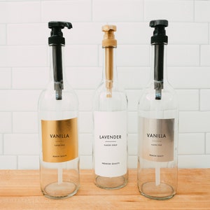 Puede incluir: Tres botellas de vidrio con tapas de bomba negras y etiquetas. Las etiquetas son doradas, blancas y plateadas. Las botellas est&aacute;n etiquetadas con "Jarabe de sabor a vainilla", "Jarabe de sabor a lavanda" y "Jarabe de sabor a vainilla".