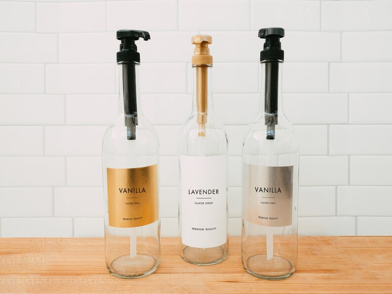 Puede incluir: Tres botellas de vidrio con tapas de bomba negras y etiquetas. Las etiquetas son doradas, blancas y plateadas. Las botellas est&aacute;n etiquetadas con "Jarabe de sabor a vainilla", "Jarabe de sabor a lavanda" y "Jarabe de sabor a vainilla".