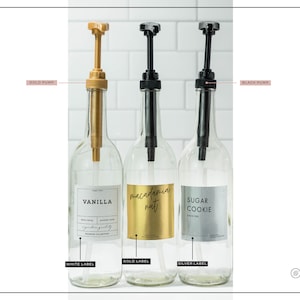 May include: Three clear glass bottles with pump dispensers. One has a gold pump and a white label that says "VANILLA". The second has a black pump and a gold label that says "macadamia nut". The third has a black pump and a silver label that says "SUGAR COOKIE".