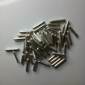 Puede incluir: Un conjunto de chispas brillantes de color plateado en forma de varilla. Estas pequeñas decoraciones de aspecto metálico están dispersas sobre una superficie blanca. Las chispas son cilíndricas con extremos redondeados, reflejando la luz.