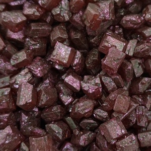 May include: Close-up of a pile of dark red, crystalline structures. The crystals have a faceted, geometric shape and a slightly iridescent sheen, reflecting light. The overall appearance suggests a collection of raw minerals or gemstones.