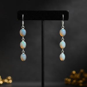 Könnte beinhalten: Ein Paar silberner Ohrhänger, jeweils mit drei ovalen Opalen. Die Opale haben eine milchig-weiße Basis mit blauen und gelben Lichtreflexen. Die Ohrringe werden auf einem schwarzen Ständer vor dunklem Hintergrund präsentiert.