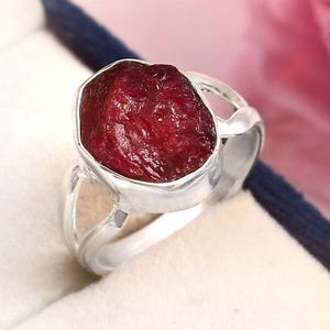 May include: A silver ring featuring a large, raw, deep red gemstone. The gemstone is irregularly shaped and set in a polished silver band. The ring is presented in a blue jewelry box, with a soft pink background.