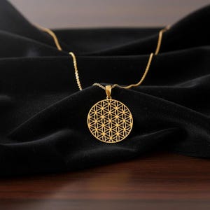 May include: A gold necklace with a circular pendant displaying a geometric flower of life pattern. The pendant is suspended from a fine chain, set against a black fabric backdrop. This is a piece of jewellery.