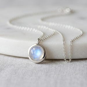 May include: A silver necklace with a round pendant featuring a blue and white iridescent stone. The pendant is set in a silver bezel and hangs from a delicate silver chain. The necklace is displayed on a white surface.