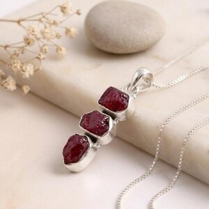 May include: A silver necklace featuring a pendant with three vertically aligned, raw, deep red garnet stones set in polished silver bezels. The pendant hangs from a delicate silver box chain, showcasing a clean, metallic finish and natural stone texture.