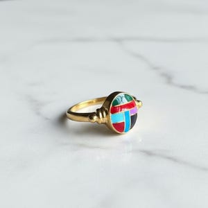 Könnte beinhalten: Ein goldener Ring mit einem farbenfrohen, geometrischen Design. Der Ring hat ein rundes, emailliertes Mittelstück mit roten, blauen, grünen und schwarzen Segmenten. Das Band ist aus einfachem, poliertem Gold. Der Ring ist vor einem weißen Hintergrund platziert.