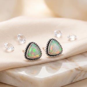 May include: A pair of sterling silver stud earrings with triangular opal stones. The opals display a play of color with green, orange, and red hues. The earrings have a twisted rope design around the edges and are set against a neutral background.