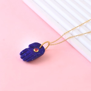 May include: A gold chain necklace with a blue lapis lazuli hamsa hand pendant featuring a red gemstone in the centre.