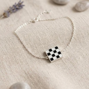 May include: A silver chain bracelet with a square charm featuring a black and white checkered pattern. The bracelet is displayed on a neutral-toned fabric, with a sprig of lavender and small stones in the background.