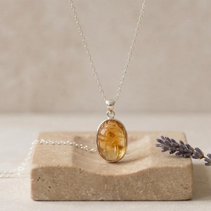 May include: A silver necklace with an oval pendant featuring a carved, amber-colored scarab beetle. The pendant hangs from a delicate chain. The necklace rests on a light brown, rectangular stone surface.