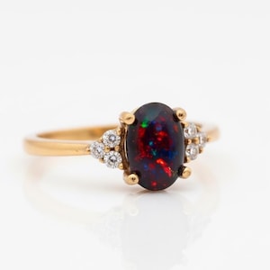 May include: A gold ring featuring an oval black opal gemstone with red and green flashes. The ring is accented with small, round diamonds on either side of the opal. The band is a simple, polished gold.