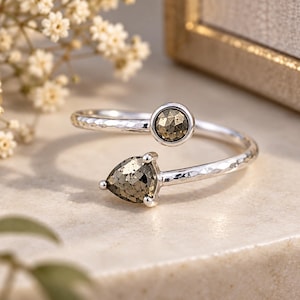 Puede incluir: Un anillo abierto de plata con una textura de banda martillada. El anillo está adornado con dos piedras metálicas facetadas: una piedra circular en la parte superior y una piedra en forma de pera en la parte inferior, ambas engastadas en garras de plata.