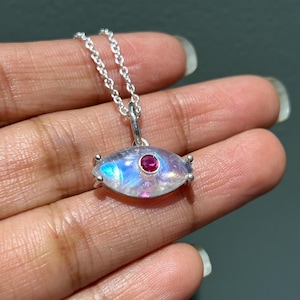 Op de afbeelding: Een zilveren ketting met een hanger van maansteen in de vorm van een oog. Het oog heeft een kleine rode edelsteen in het midden.