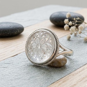 May include: A silver ring with a large, circular face showcasing a detailed floral pattern. The ring's face is framed by a twisted rope-style border. The ring rests on a grey surface with small stones and sprigs of white flowers.