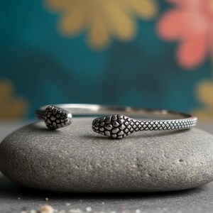 May include: A silver-colored snake bracelet with textured scales and open ends. The bracelet is resting on a gray stone, with a blurred background of teal, yellow, and pink.