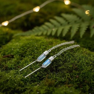 Op de afbeelding: Zilveren oorbellen met een lange, dunne staaf en een kleine, heldere kristallen hanger met een blauwe tint. De oorbellen worden gepresenteerd op groen mos, met feeërieke lichtjes en een varen op de achtergrond.