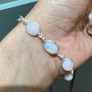 以下が含まれることがあります： 4つの楕円形の白いオパールがついたシルバーのブレスレット。ブレスレットは人の手首につけています。