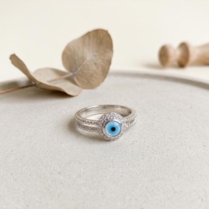 May include: A silver ring featuring a central blue eye design, encircled by small, clear stones. The ring has a double band. The eye has a light blue iris and a dark blue pupil. The ring is displayed on a light grey surface.