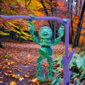 Puede incluir: Una figurita de perezoso verde articulada colgada de una rama morada. El perezoso tiene un patrón texturizado de hojas y está posado con los brazos levantados por encima de su cabeza. El fondo es una imagen borrosa de un bosque de otoño.