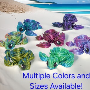 May include: A collection of seven colorful, 3D printed turtle figurines, each with a rose-shaped shell. The turtles are arranged on a white sandy beach background. Multiple Colors and Sizes Available!