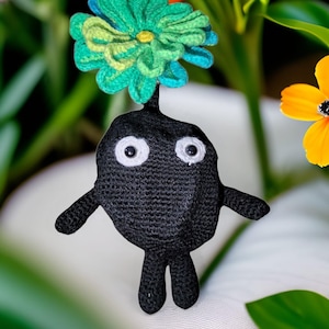 May include: A handmade black crochet figure with white eyes and small arms and legs. A large, colorful flower in shades of green and blue sits atop the figure's head. The figure is set against a blurred background of greenery and flowers.