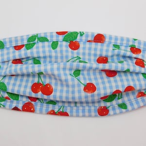 Op de afbeelding: Een blauw en wit gingham stoffen mondkapje met een rode kersenprint. Het masker heeft plooien en elastische oorlussen.