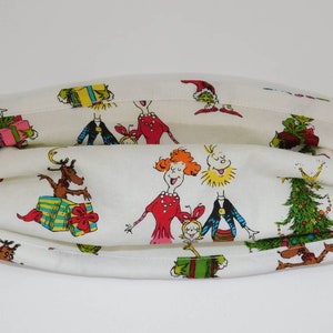 May include: White fabric face mask with a Christmas theme featuring characters from the book "How the Grinch Stole Christmas!" The characters include the Grinch, Cindy Lou Who, and the Grinch's dog Max. The mask has a pleated design and is made of 100% cotton.