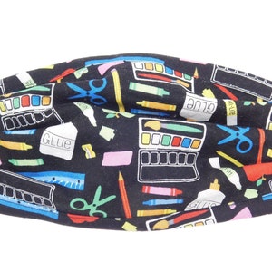 Puede incluir: Máscara facial de tela negra con un estampado colorido de útiles escolares, incluyendo tijeras, pegamento, lápices de colores y pinceles.