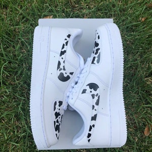 May include: White leather low-top sneakers with a black and white cow print design. The Nike swoosh logo is also in the cow print design.