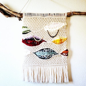 May include: A woven wall hanging with a natural beige background and colorful geometric shapes. The shapes are woven with different colored yarns and include red, yellow, blue, green, and white. The hanging is attached to a natural branch.